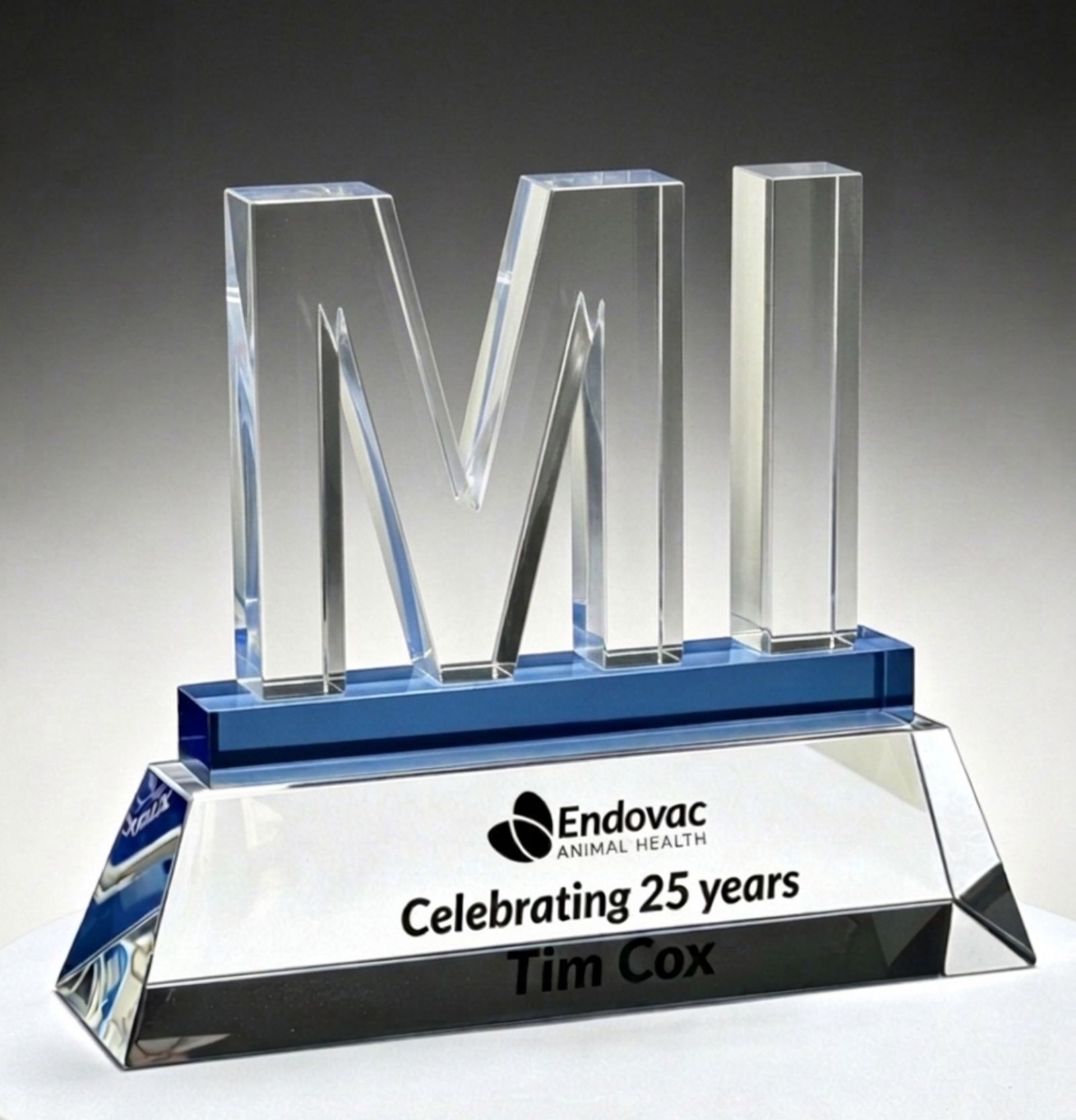 Clear crystal service award with large letters “MI” rising from a blue accent strip, standing on a beveled crystal base. The base is printed with the Endovac Animal Health logo, “Celebrating 25 years,” and the name Tim Cox, photographed against a dark background.